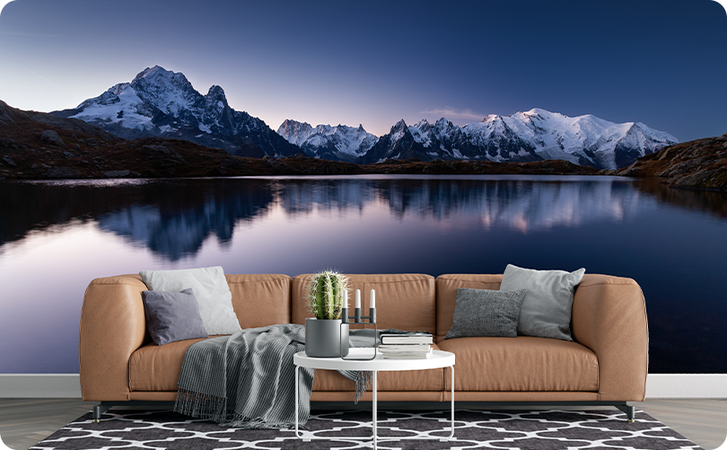 Twilight mountain lake wallpaper mural with snowy peaks reflected in blue water behind a tan sofa